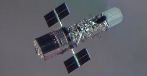 A close-up view of a space telescope with solar panels extended on either side, floating against the backdrop of space with a faintly visible planet or atmosphere.