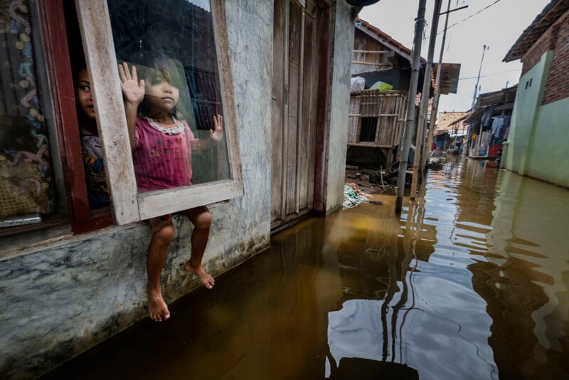 Two children look out from a window above floodwaters in a narrow alley; one child sits on the window ledge with feet above the water, while the other stands behind the glass. Flooded street and houses are visible outside.