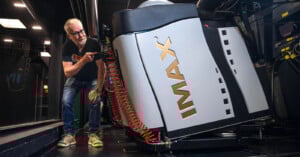 A man with glasses and a gray beard inspects a large, tilted IMAX projector in a dark room, smiling and pointing at the machine.