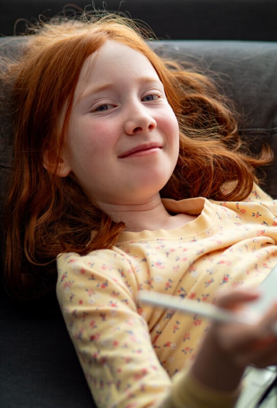 A young girl with long red hair lies on a couch, smiling softly. She wears a yellow shirt with a small floral pattern and holds a white object in her hand, possibly a tablet or notebook.