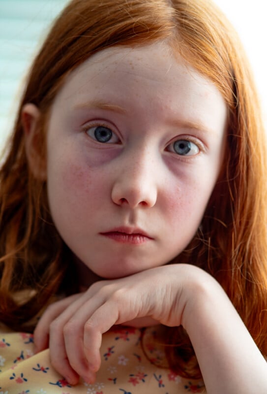 A young girl with long red hair and blue eyes gazes directly at the camera, her expression serious. She rests her chin on her hand and wears a light-colored, floral-patterned top. Soft light illuminates her face.