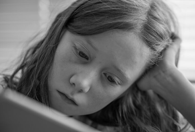 A young girl with long hair rests her head on her hand and looks intently at a book or notebook in front of her. The image is in black and white, capturing a thoughtful, focused expression.