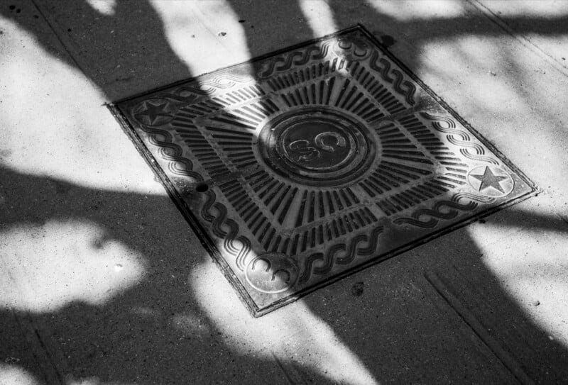 A decorative metal utility cover set in a sidewalk, featuring geometric patterns, stars, and the letters "SG" in the center, with tree shadows cast across its surface.