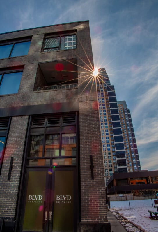 A tall brick building with large windows and “BLVD BELTLINE” on the doors, stands in front of modern high-rises. The sun creates a starburst effect at the building's corner; snow covers the ground.