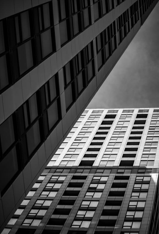 Black and white photo of tall modern buildings shot from below, creating dramatic lines and geometric patterns as they rise toward the sky, with windows and shadows accentuating the architectural design.