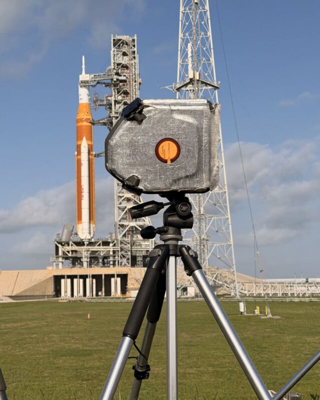 A camera wrapped in a protective covering is mounted on a tripod in the foreground, with a large orange and white rocket on a launch pad and a tall metal structure visible in the background under a blue sky.
