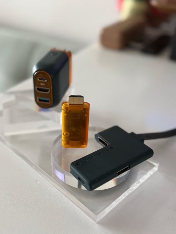 Three electronic adapters, including two black USB hubs and one small orange HDMI adapter, are displayed on a clear acrylic stand on a white surface. The background is softly blurred.