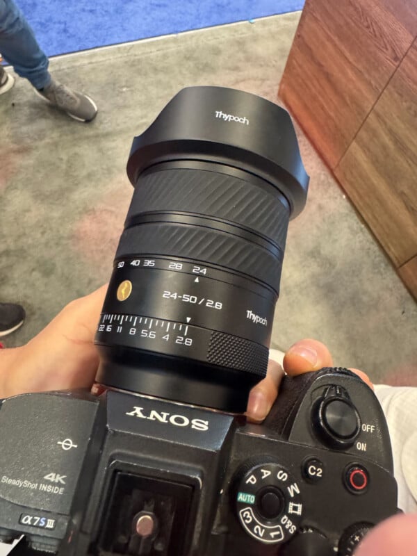 Close-up of a person holding a Sony camera with a Thypoch 24-50mm f/2.8 lens attached. The camera is pointed downward, and the surrounding environment includes a carpeted floor and wooden surface.