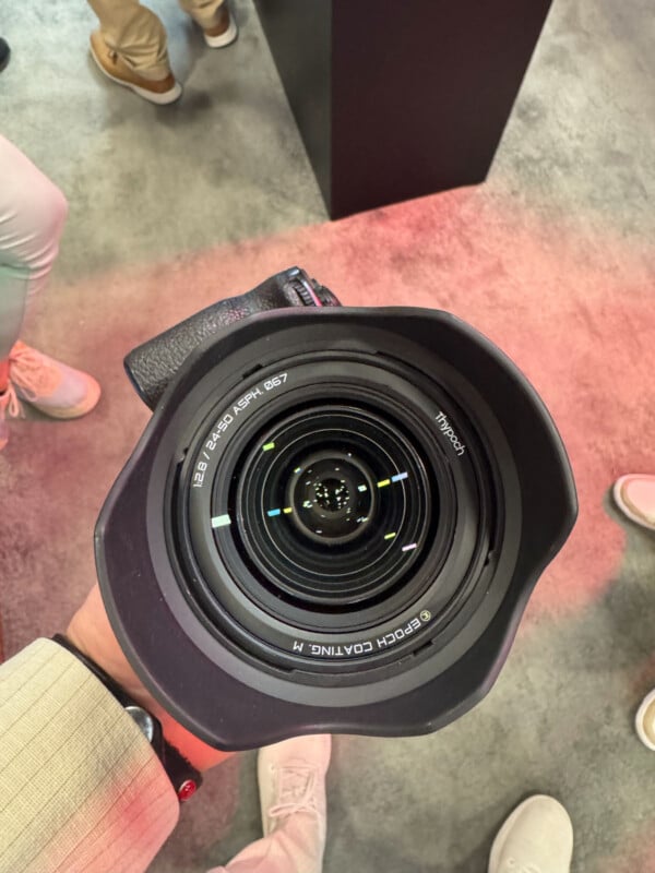 A person holds a large camera with a wide-angle lens, pointed upward. The lens has visible markings and reflections. Several people wearing light-colored shoes stand on a gray and pinkish floor in the background.