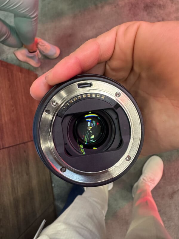 A hand holds a camera lens facing upward, showing the electronic contacts and glass elements. A person wearing light shoes and pants stands on a textured floor in the background.