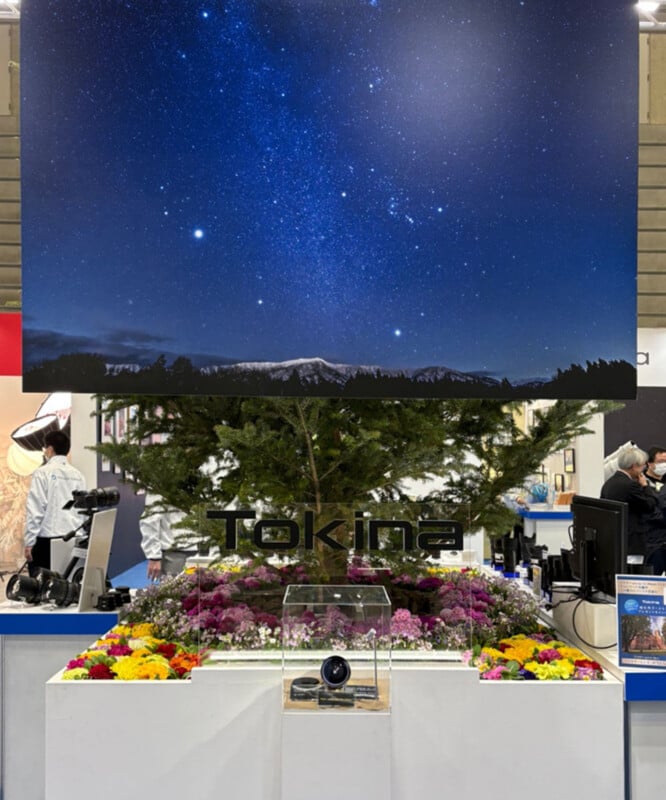 Un stand de exhibición equipado con una lente de cámara Tokina, rodeado de flores coloridas y árboles verdes, con grandes fotografías del cielo estrellado y vistas de las montañas sobre el stand. Al fondo se ve claramente a la gente.