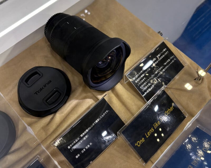 Una superficie marrón que muestra una lente de cámara gran angular Tokina y su tapa, con un cartel informativo al lado que dice "¡Una lente única!" Texto amarillo.