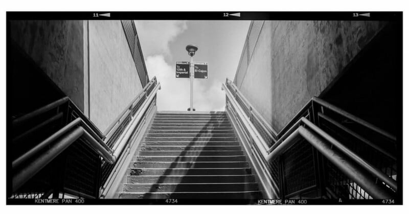 A black-and-white photo of outdoor stairs leading up between two walls, with handrails on both sides. At the top, a signpost and a lamp are visible against a partly cloudy sky.