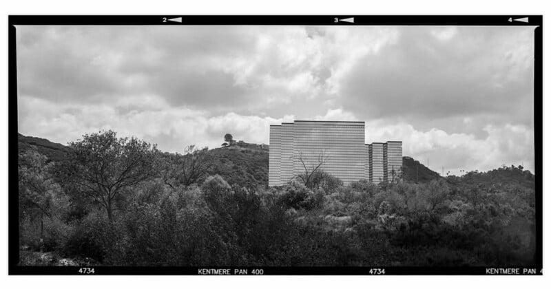 A modern, rectangular building stands on a hillside surrounded by dense trees and shrubbery under a cloudy sky, captured in black and white with a film photo border.