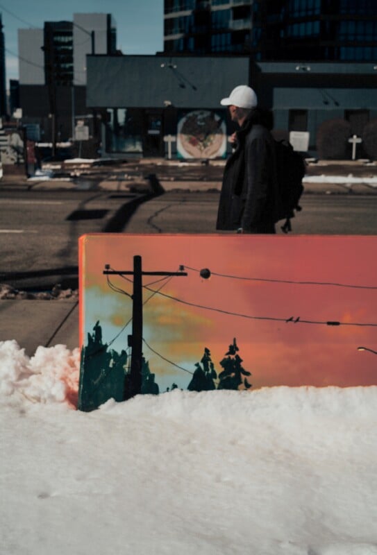 A person in a white cap walks on a snowy city street. In the foreground, a painted utility box shows power lines and trees against an orange sky. Snow covers the ground and urban buildings are in the background.
