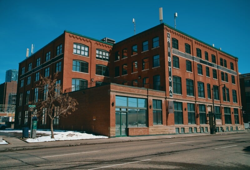 A large brick industrial-style building with large windows, a sign reading "TRIBUTOR" on the corner, and some snow on the ground, under a clear blue sky.