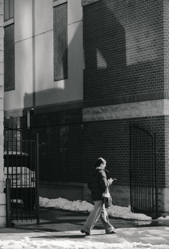 A person wearing a winter coat walks past a brick building and open iron gate on a snowy sidewalk, while looking down at their phone. Shadows and sunlight create patterns on the building’s wall.