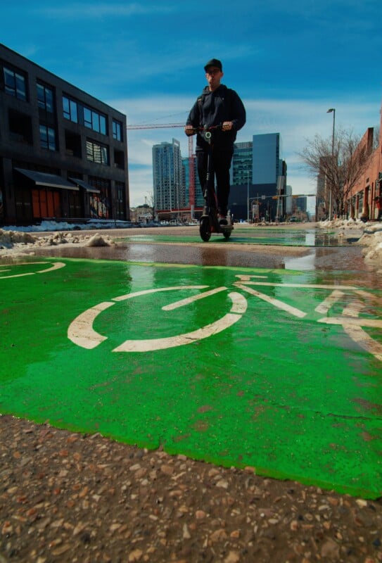 A person rides an electric scooter on a wet, green bike lane through a city. Buildings and melting snow are visible on either side, with a partly cloudy sky above.