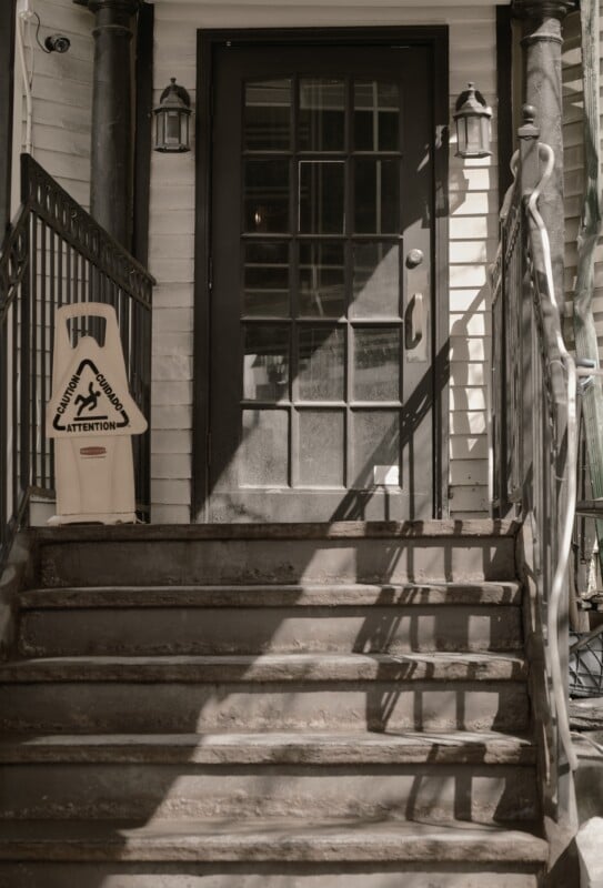 A black metal staircase leads up to a glass-paneled door with two lantern-style lights on either side. A yellow caution sign stands at the top of the stairs, warning of a wet floor.