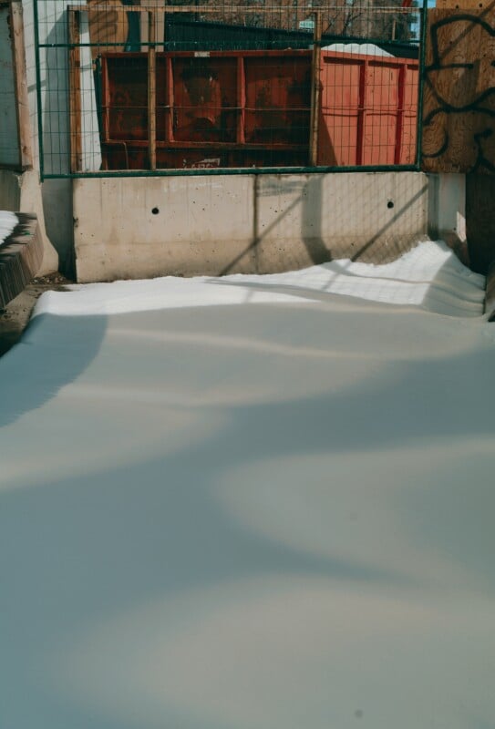 A construction site with a layer of snow covering the ground, surrounded by concrete walls, fencing, and red metal containers in the background. Shadows are cast across the snow.