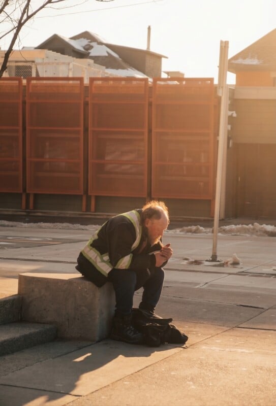 A man wearing a reflective safety vest sits on a concrete bench outdoors, hunched over and eating food from his hands, with a warm sunlight casting shadows and snow visible in the background.