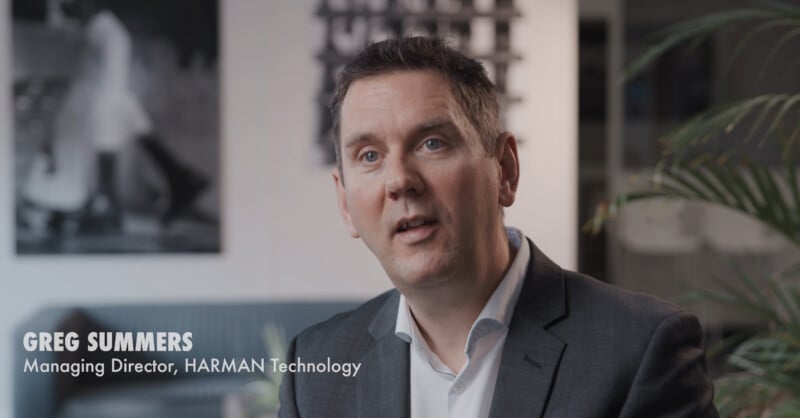 A man in a suit sits indoors, looking slightly to the side. Text on screen reads "Greg Summers, Managing Director, HARMAN Technology." A blurred black-and-white photo and green plant are in the background.