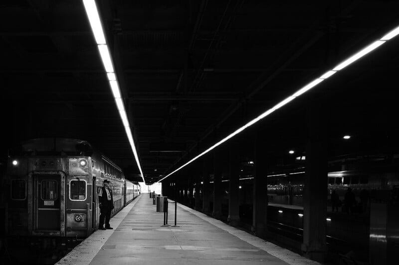 A black and white photo of a dimly lit train station platform with a train on the left side. A person stands near the train, while bright overhead lights lead toward the vanishing point in the distance.