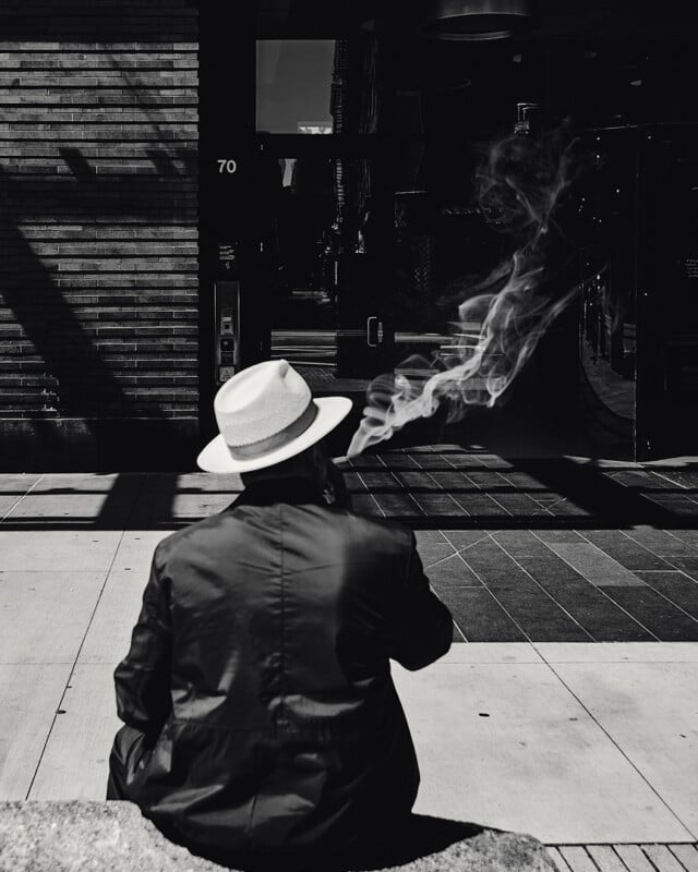 A person wearing a light-colored hat and dark coat sits on a city sidewalk, facing away, exhaling a large plume of smoke that swirls in the air. The image is in black and white, with sharp shadows and urban surroundings.