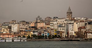 A waterfront cityscape with densely packed buildings in various colors and the tall, historic Galata Tower standing prominently among them under a cloudy sky in Istanbul, Turkey. Boats are docked along the shore.