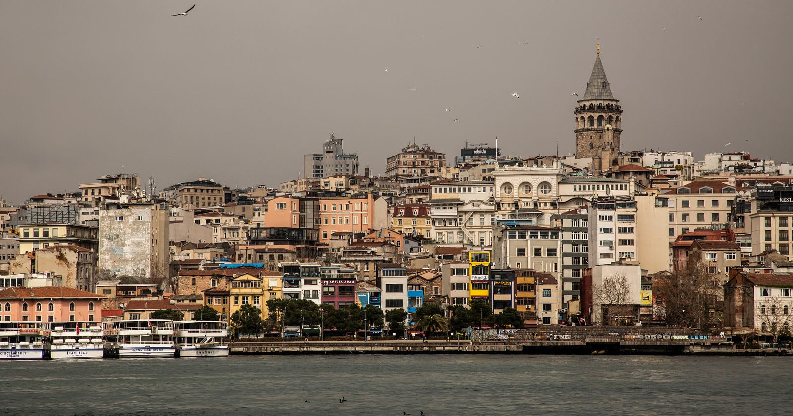A waterfront cityscape with densely packed buildings in various colors and the tall, historic Galata Tower standing prominently among them under a cloudy sky in Istanbul, Turkey. Boats are docked along the shore.