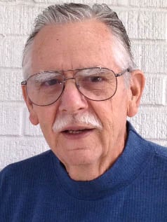 An older man with gray hair, a mustache, and glasses, wearing a blue sweater, stands in front of a white brick wall.