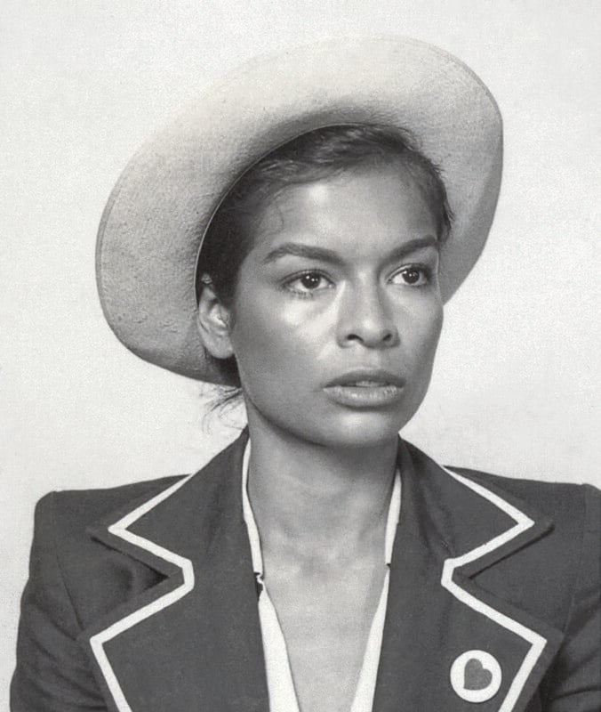 A woman with a serious expression wears a light-colored hat and a dark blazer with light trim. The image has a vintage, black-and-white style.