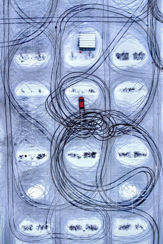 Aerial view of a red car in a snowy parking lot with circular tire tracks surrounding it; a small structure and scattered bushes are visible among the parking spaces.