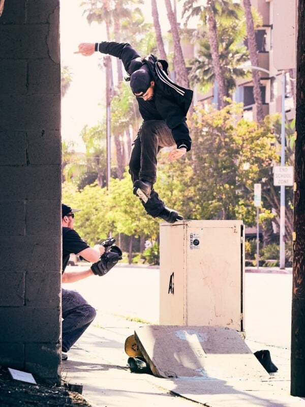 A skateboarder performs a trick over a utility box on a city sidewalk while a cameraman crouches nearby, capturing the action. Palm trees and sunlit buildings are visible in the background.