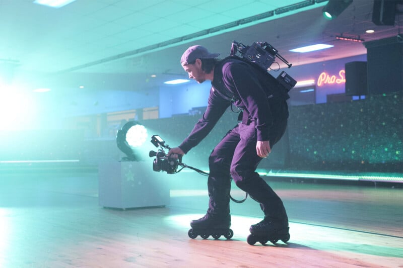 A person wearing rollerblades and a backwards cap films with a video camera and wears a camera rig backpack in a brightly lit roller skating rink. Neon lights illuminate the background.