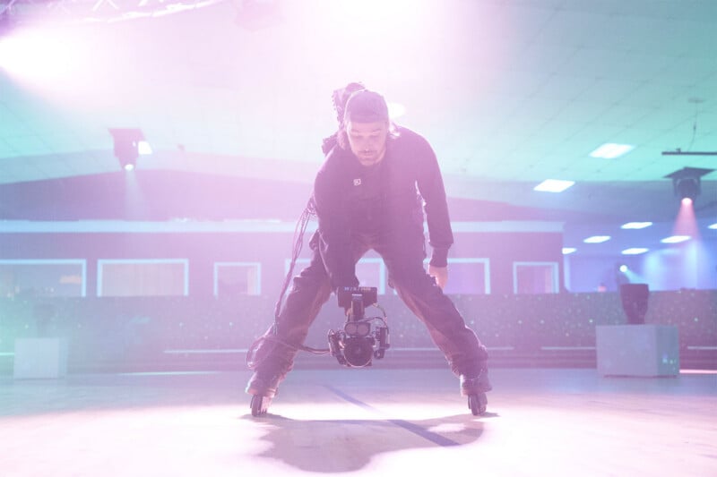 A person wearing rollerblades and a backwards cap holds a professional video camera low to the ground while skating inside a brightly lit, hazy roller rink.