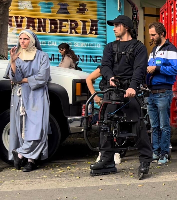 A person dressed as a nun smokes a cigarette while standing next to a white truck. Nearby, a camera operator holds film equipment, and another person looks at their phone. A colorful storefront is in the background.