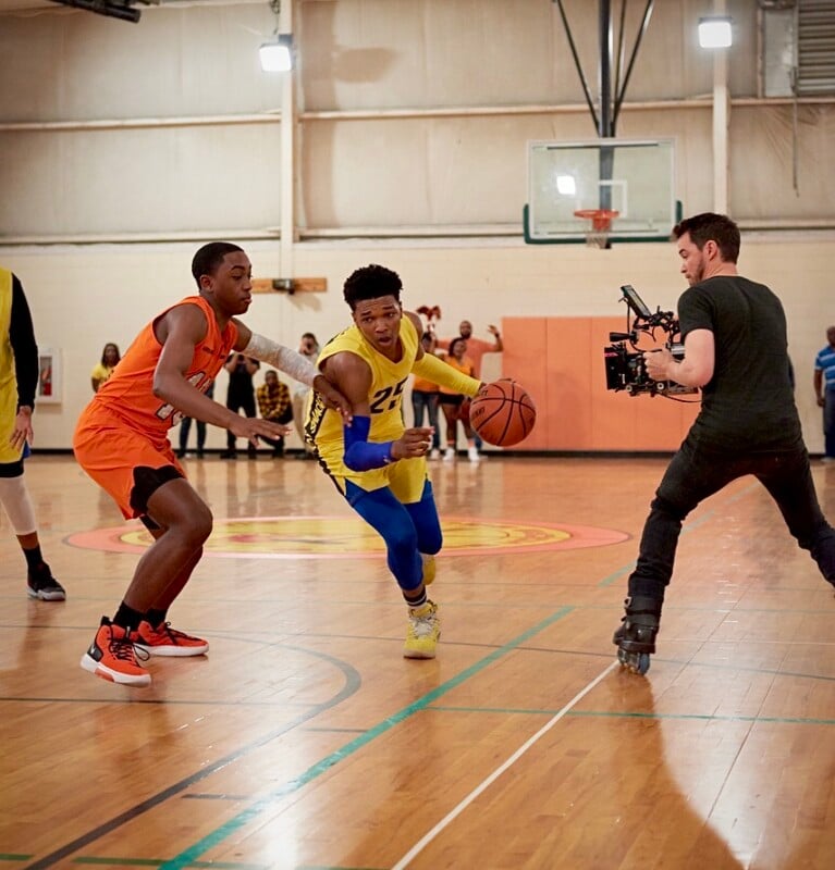A basketball player in a yellow and blue uniform dribbles past a defender in orange while a cameraman on roller skates films them on an indoor court. Spectators watch in the background.