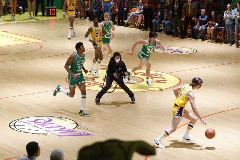 A basketball game scene with players in green Celtics and yellow Lakers uniforms in action, as a cameraman films on the court and spectators watch from the stands.