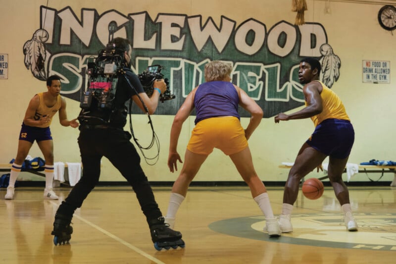 A cameraman on roller skates films two basketball players in action on a gym court, with "INGLEWOOD SENTINEL" painted on the wall in the background. Another player watches from the sidelines.