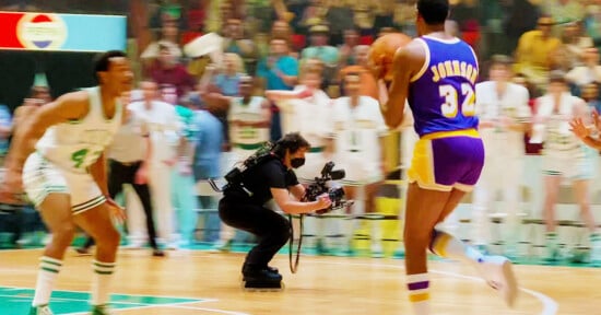 A basketball player in a purple and gold jersey with "32" on the back dribbles the ball while guarded by a player in a white and green uniform, as a cameraman films the action on a crowded court.