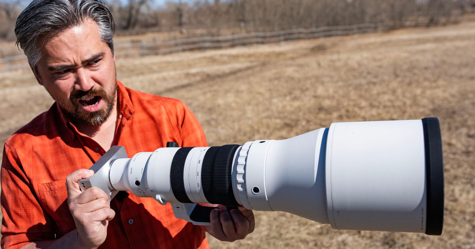 A man in an orange shirt holds a large white telephoto camera lens outdoors in a dry, grassy field, looking surprised or amazed as he examines the lens.