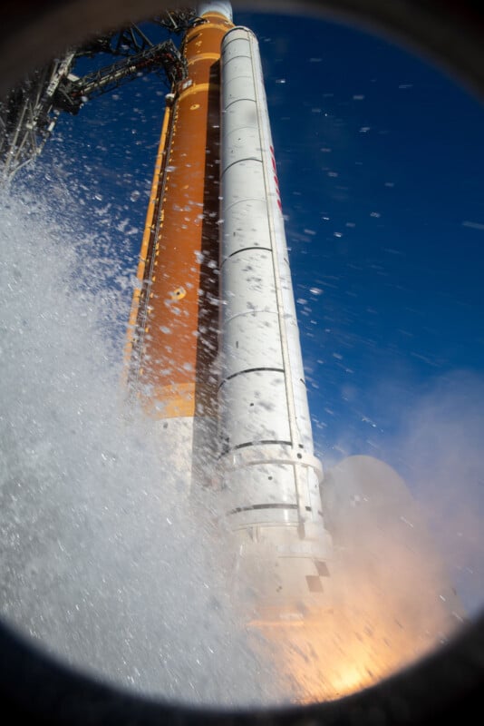 Close-up view of a rocket launching, with water spray and flames at the base, a tall orange and white rocket body rising against a clear blue sky, framed by a dark circular border.