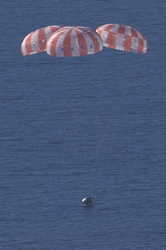 A capsule descends toward the ocean, supported by three large orange and white parachutes, with rippling blue water visible below.