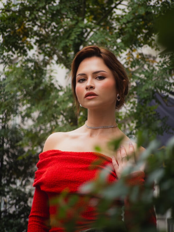 A woman with short brown hair wears a red off-the-shoulder top and a silver necklace, standing outdoors surrounded by green foliage, looking confidently at the camera.