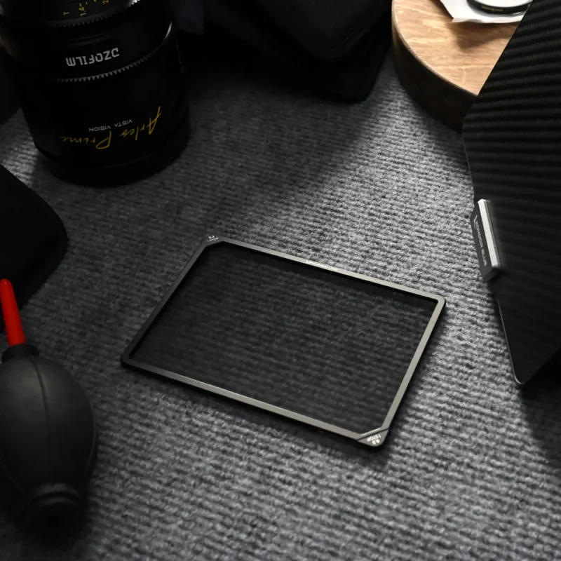 A square camera lens filter lies on a textured grey surface, surrounded by photography equipment including a camera lens, an air blower, and a round wooden item.