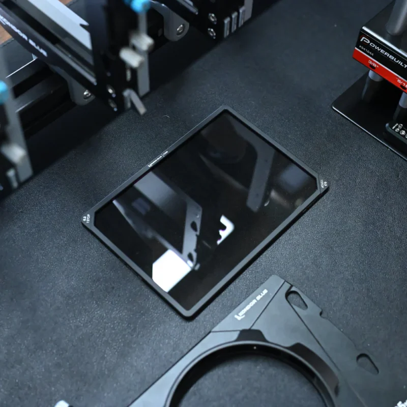 A rectangular glass panel lies flat on a dark surface surrounded by metal and mechanical components, likely part of a 3D printer or precision machine assembly. The panel reflects parts of the equipment above.