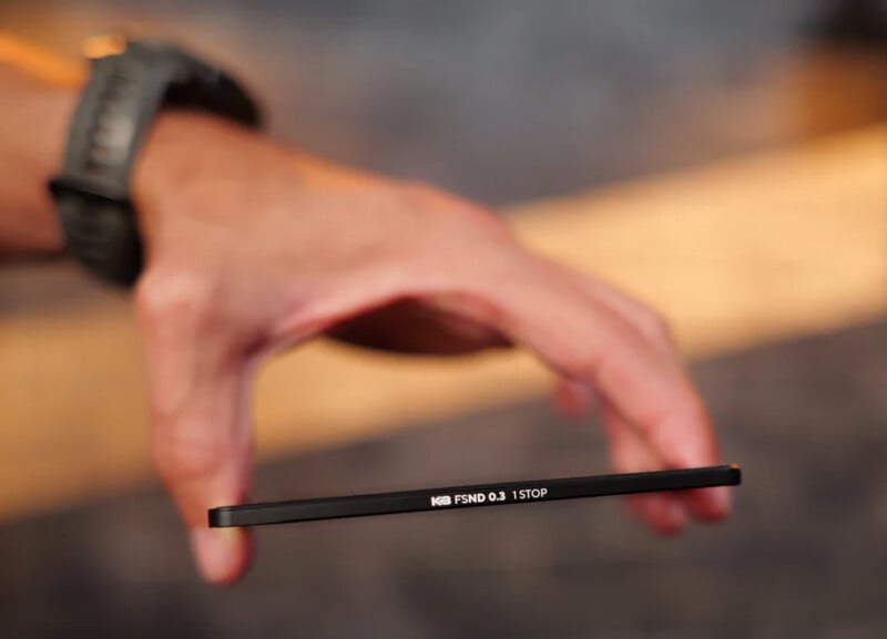 A hand with a watch holds a thin, round camera filter marked "K&F FSND 0.3 1 STOP" between the thumb and index finger, with a blurred background.