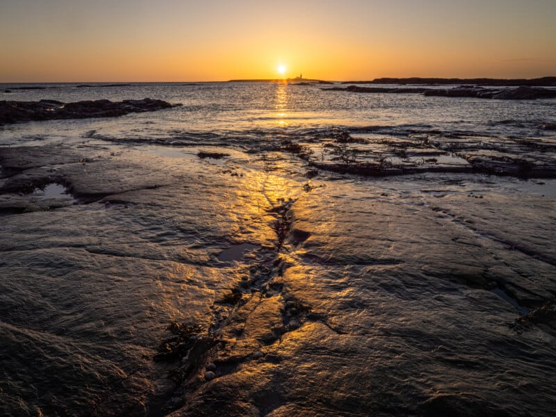 The sun sets over the ocean, casting a golden reflection across wet, rocky shorelines. The sky glows orange, and gentle waves approach the coast, creating a serene and peaceful scene.