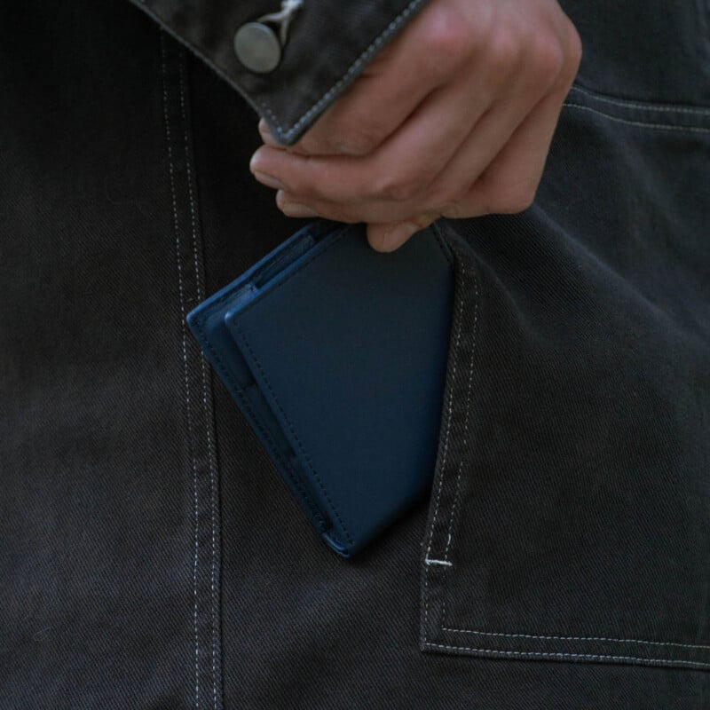 A person’s hand is shown placing or removing a slim blue wallet from the front pocket of a dark-colored jacket.